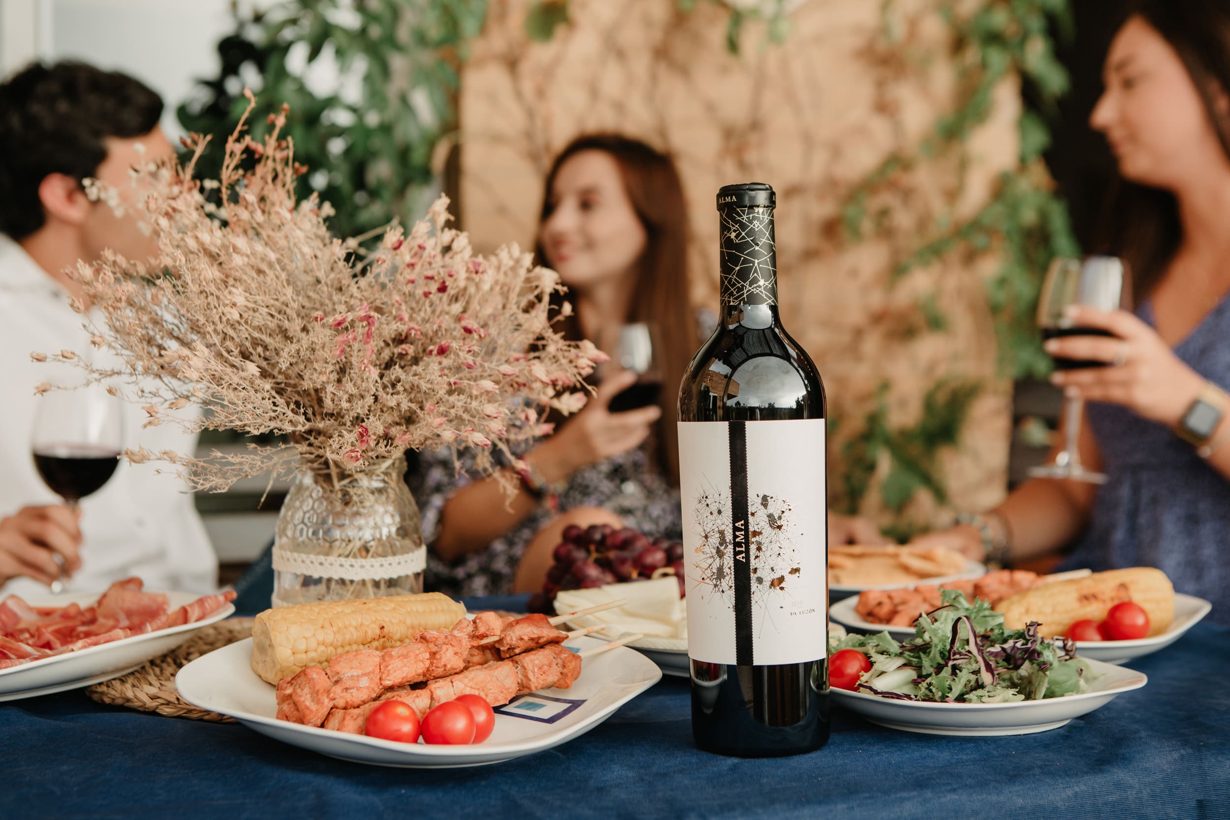 A nice table, with a variety of food and a bottle of Alma de luzon in the middle. in the background there are 3 people out of focus having a good time, wine glasses in their hands. 